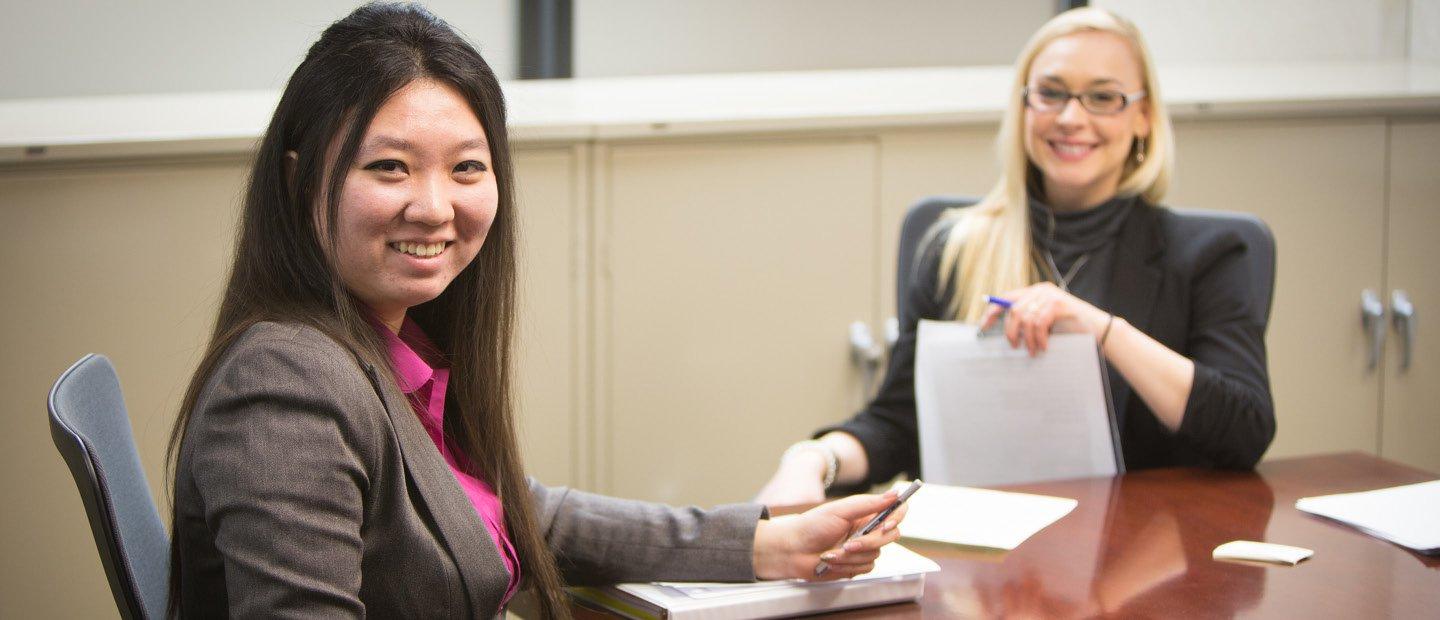 Two women seated a a table with notebooks and pens, smiling at the camera.